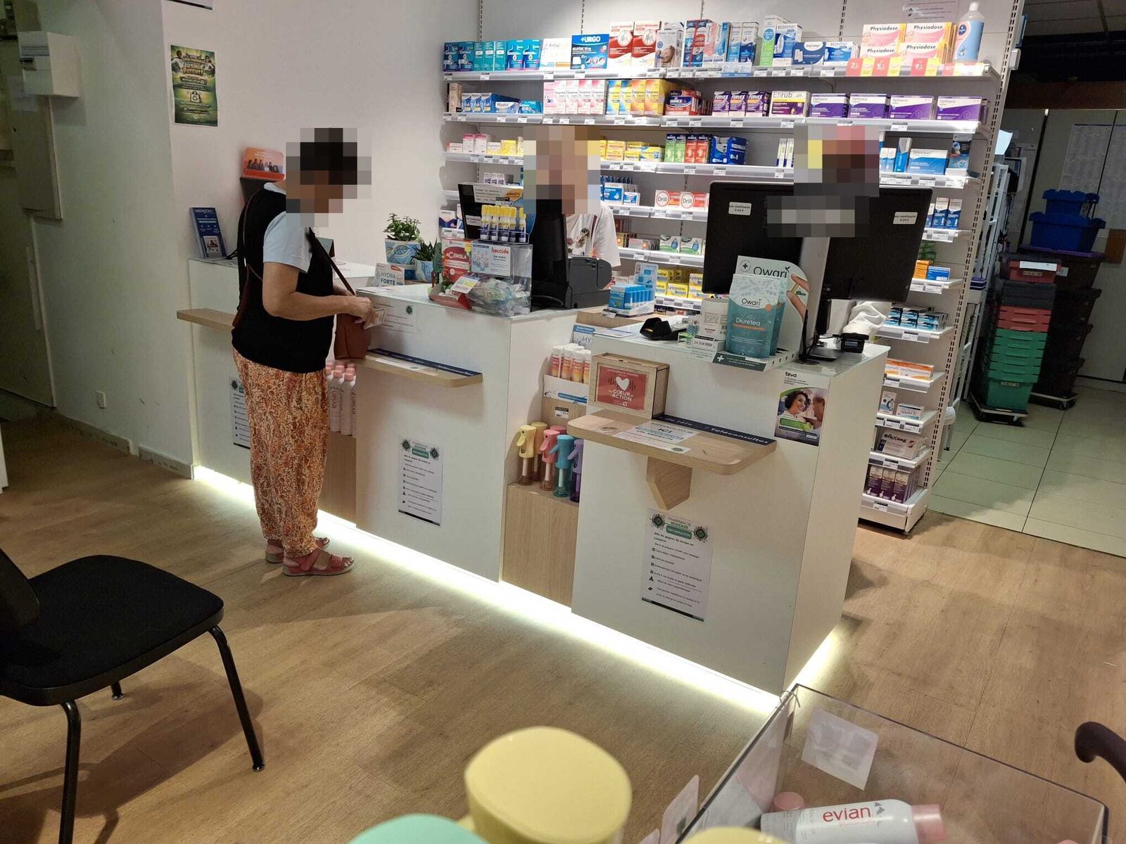 Vue d'ensemble du comptoir de la Pharmacie Moderne : une patiente est servie, deux membres de l'équipe se tiennent derrière le plan de travail, les visages sont masqués.